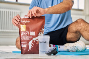 Bodylab Whey 100 -proteiinijauhepussi ja shaker treenialustalla istuvan henkilön edessä.